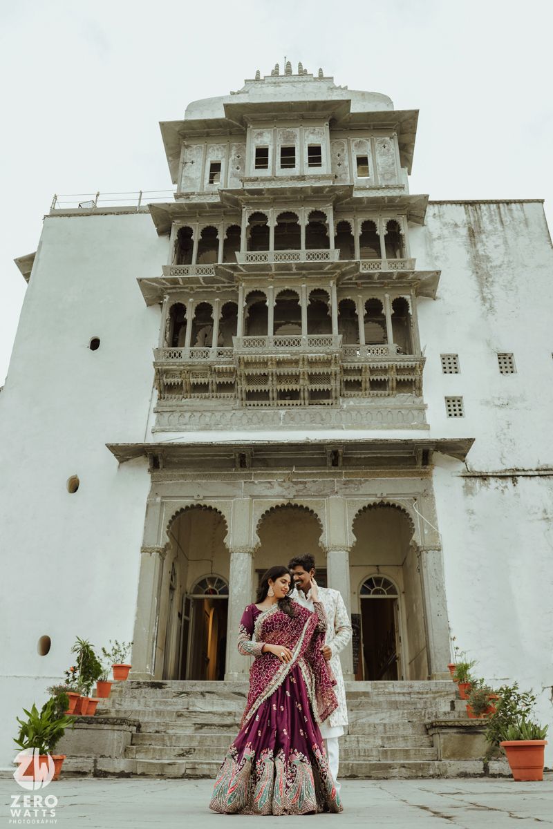 Elegant couple in traditional Indian wedding attire standing in front of white palace with ornate balconies.
