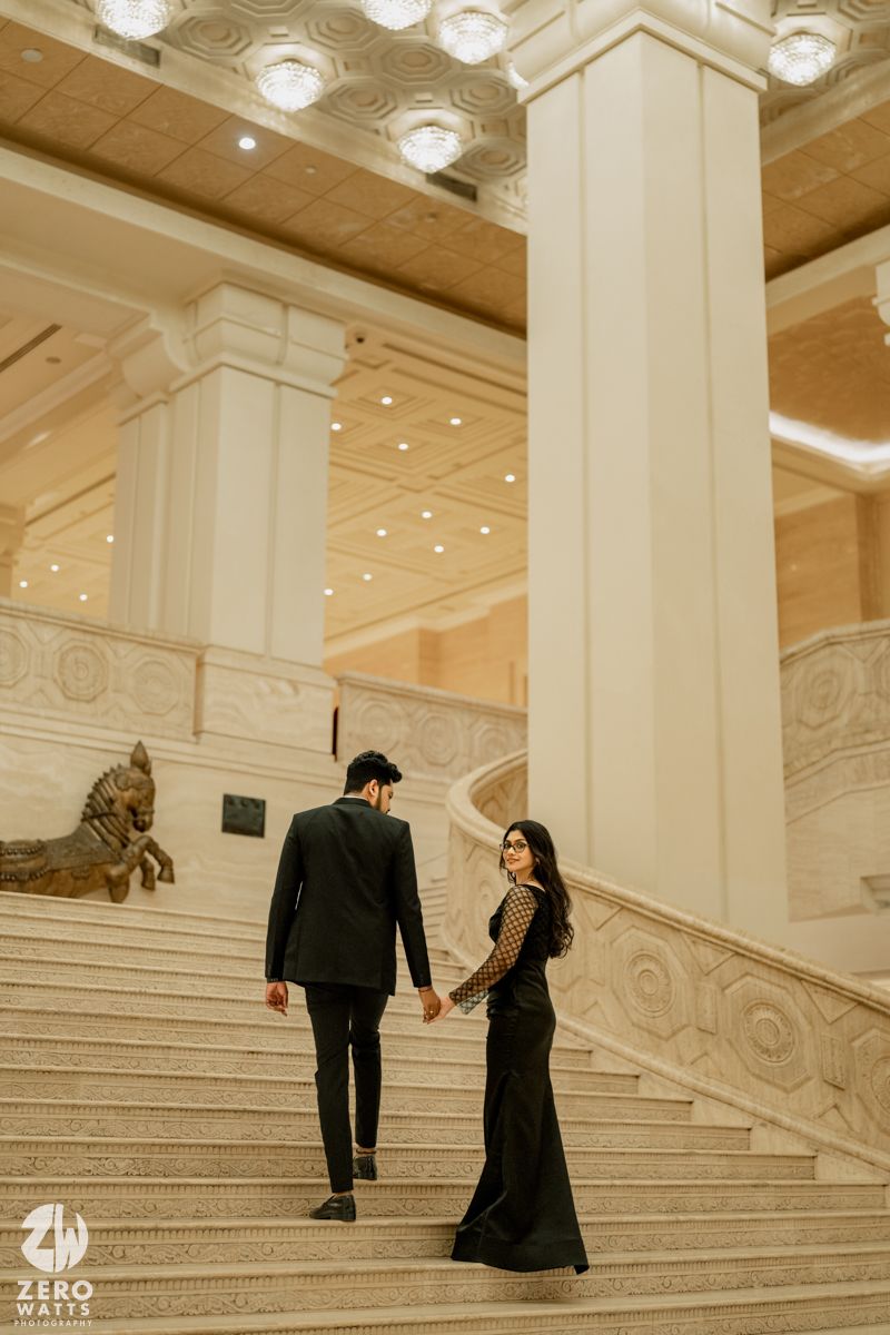 Couple in formal attire ascending grand marble staircase in elegant interior with ornate architectural details.