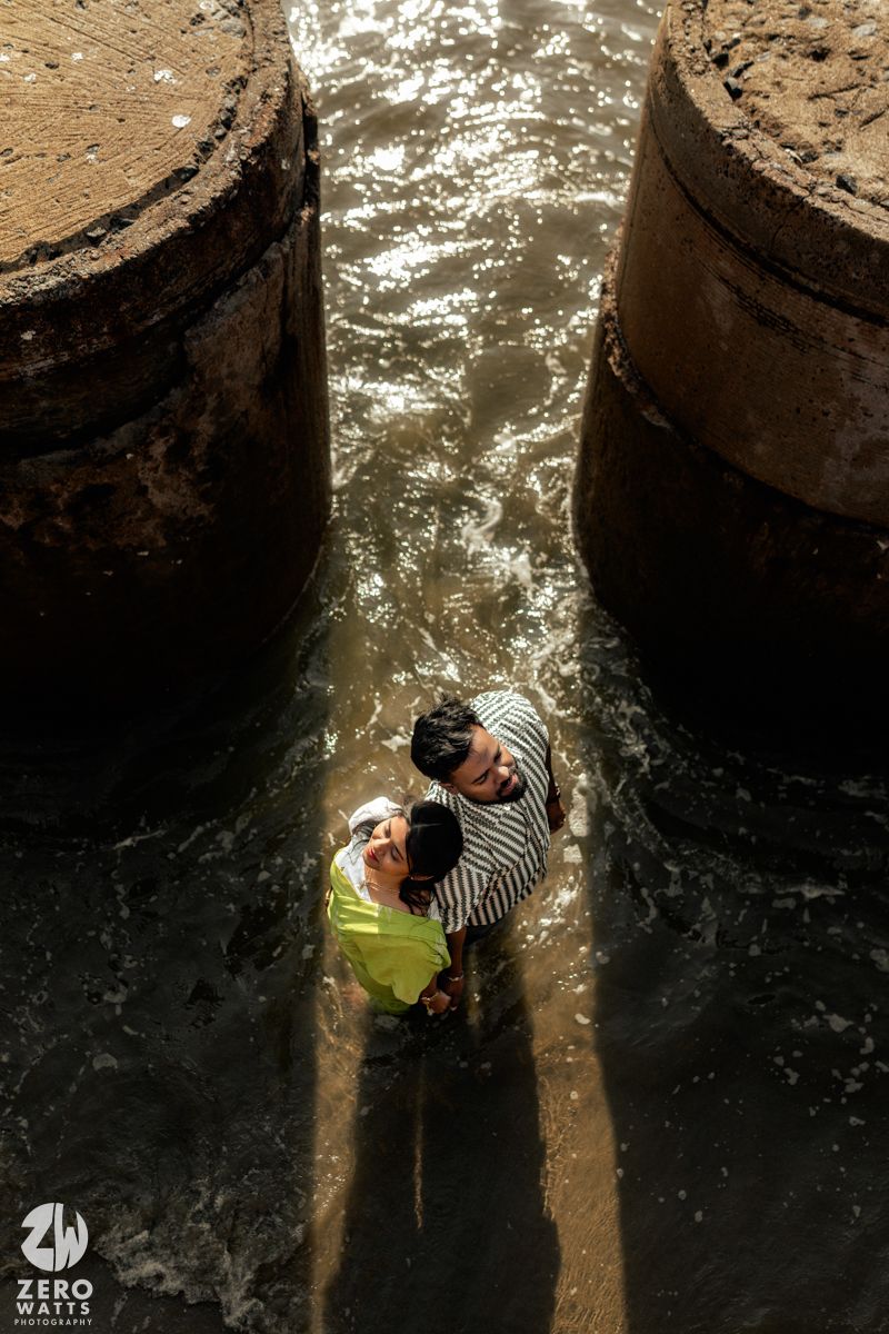 A creative overhead shot of a couple standing between dark concrete pillars near water.