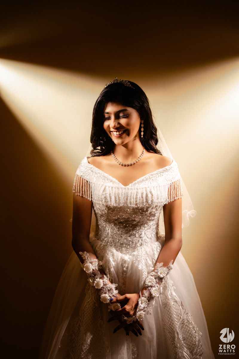 Bride in white off-shoulder wedding gown with fringe details and floral gloves, posing under golden spotlight.