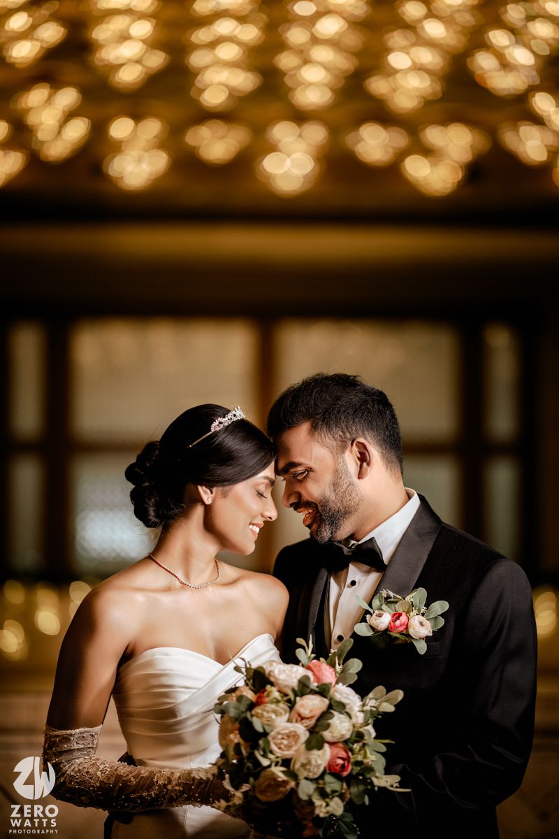 Bride and groom touching foreheads romantically, she holds a floral bouquet, glowing bokeh lights above in elegant venue.