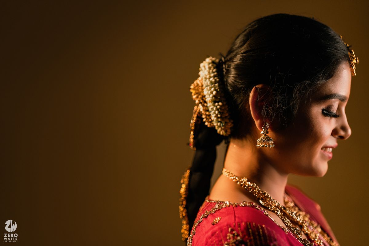 South Indian bride in profile view wearing red silk saree with gold jewelry and floral hair accessories.
