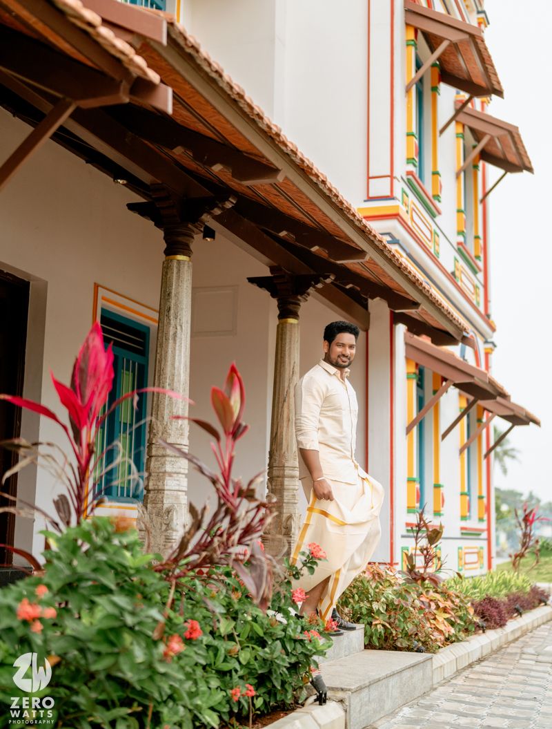Groom in white kurta and dhoti walking along a garden path beside a colorfully decorated traditional Indian building.