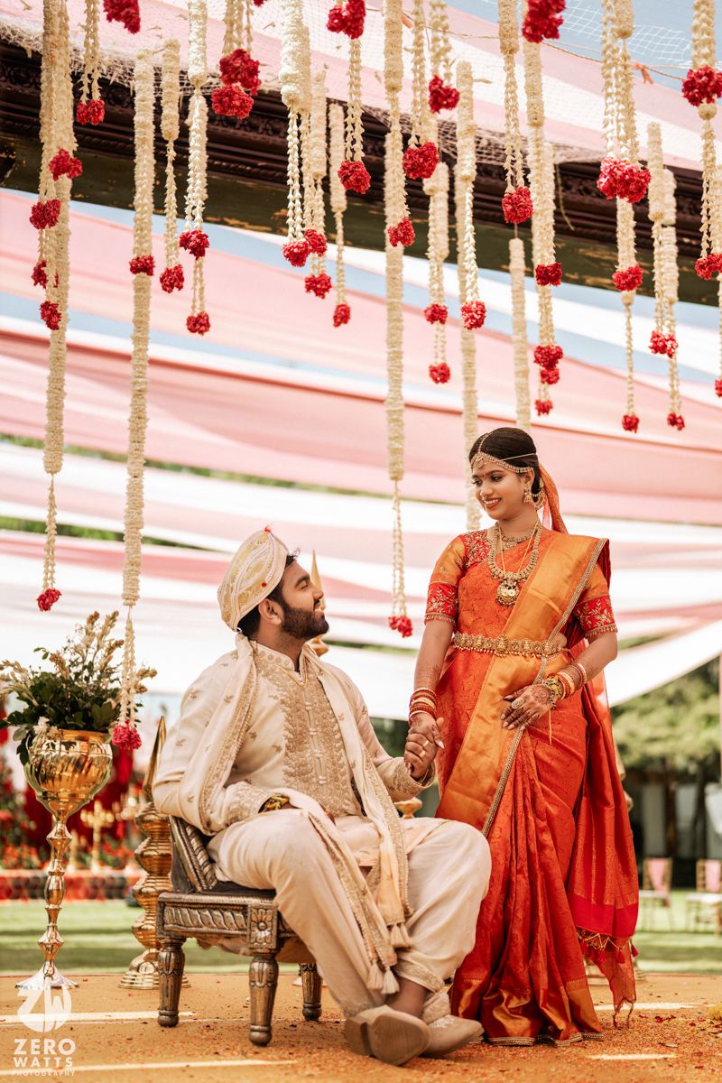 Indian bride in orange saree and groom in cream sherwani hold hands at traditional Hindu wedding ceremony.