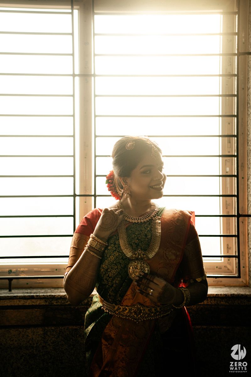 Indian bride in traditional saree and silver jewelry poses by sunlit window in elegant bridal portrait.