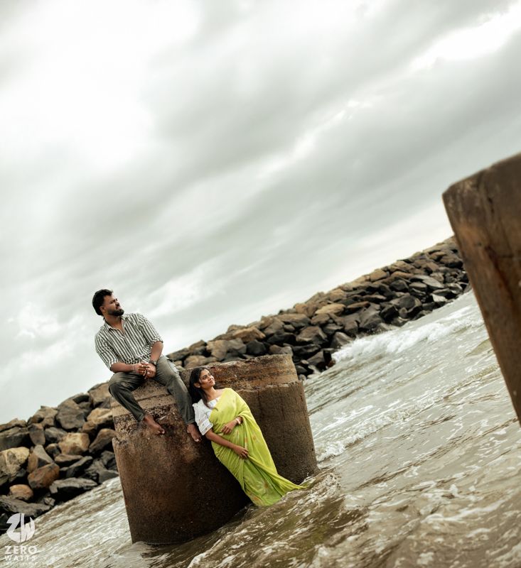 Couple poses near concrete pillars on a rocky beach during overcast weather.