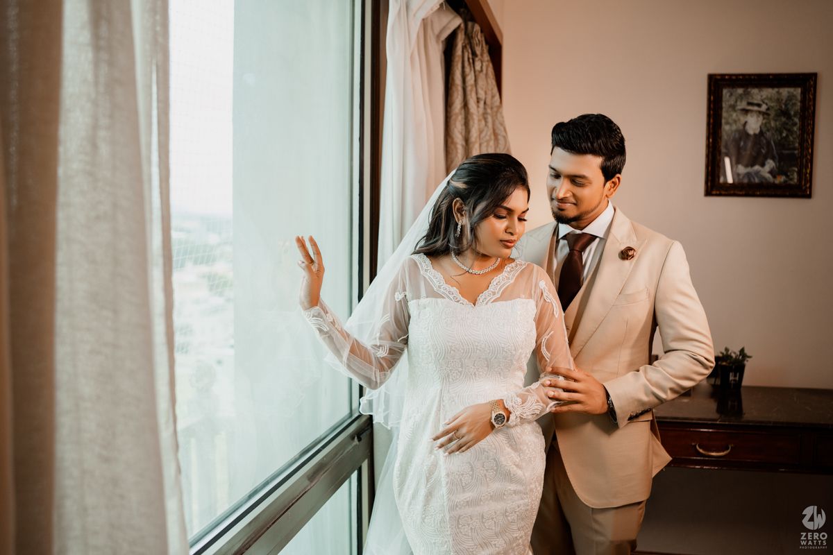 A romantic wedding photoshoot featuring a couple in white and beige attire by a window with soft lighting.