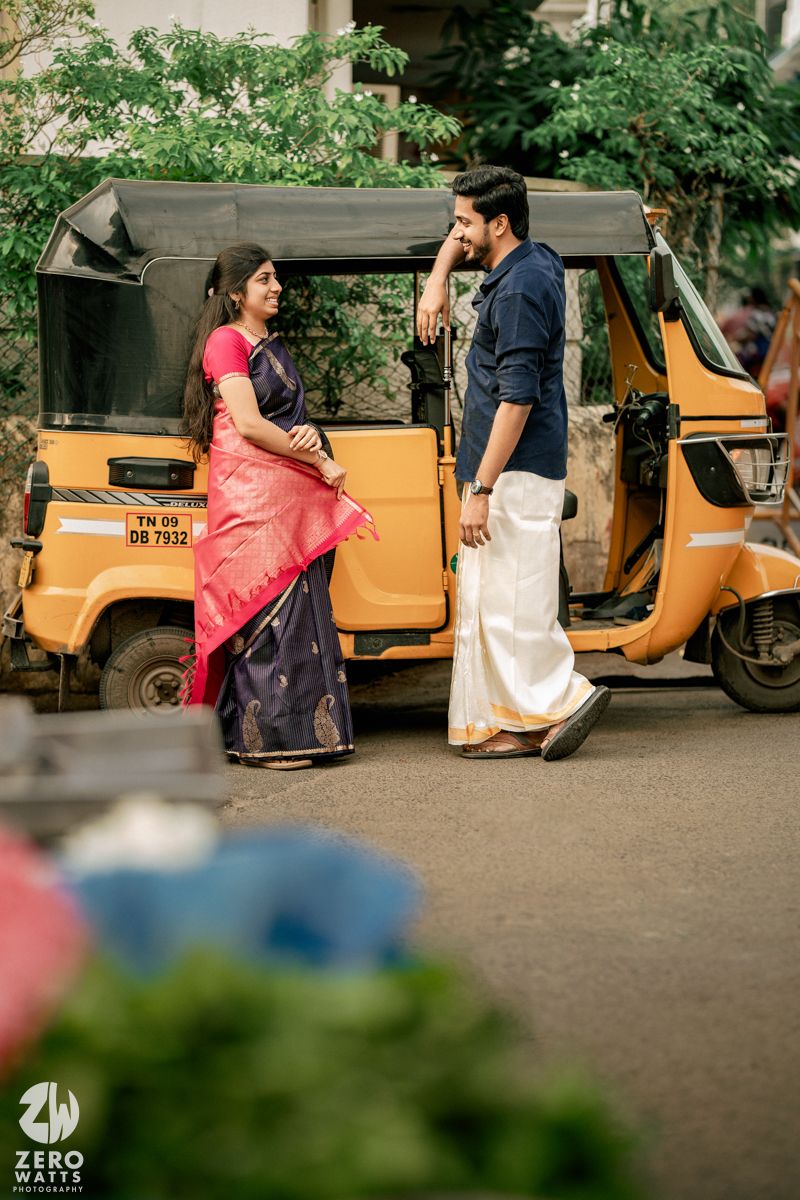 Couple interacting playfully near a parked yellow auto rickshaw on a street with greenery in the background.