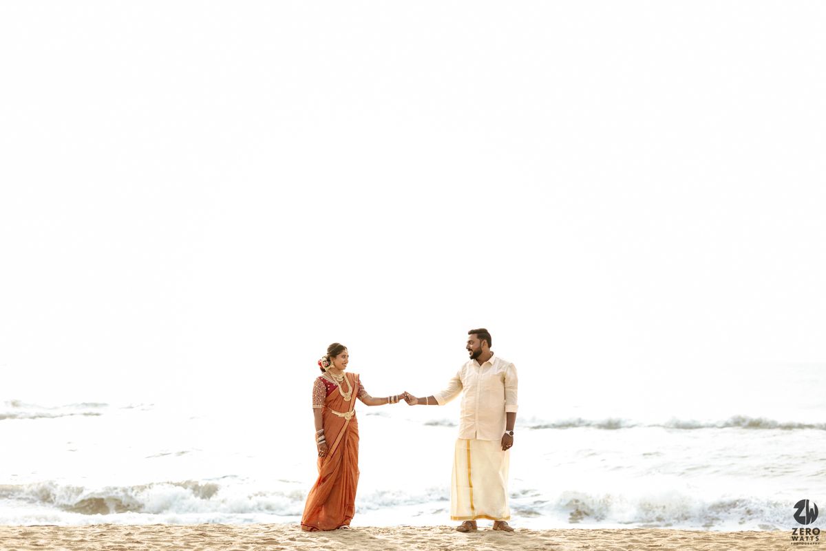 A couple in traditional Indian attire holds hands while walking along a sandy beach with ocean waves in the background.