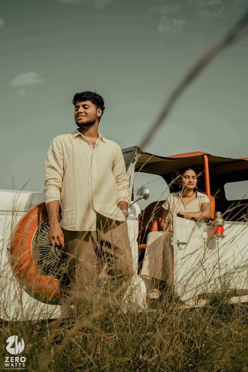 Vintage-inspired photoshoot featuring a classic car and figures in light clothing against a moody sky and tall grass.