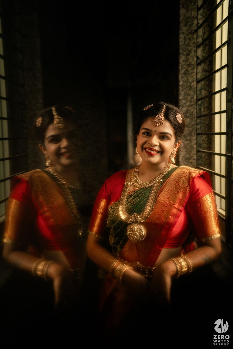 A smiling South Indian bride in red and green silk saree with gold jewelry, reflected in a mirror during wedding preparations.