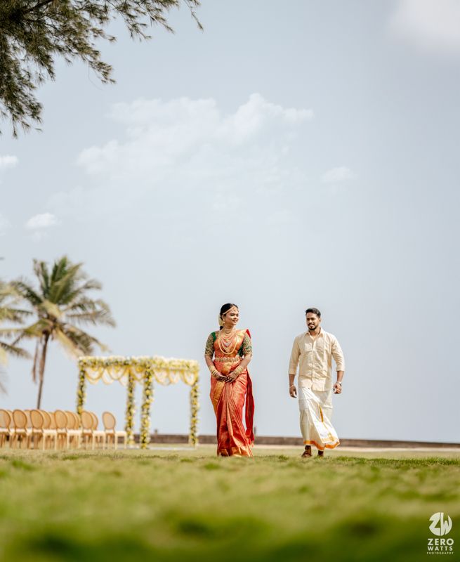 Couple in traditional Indian wedding attire walking on green lawn with floral mandap and palm trees backdrop.