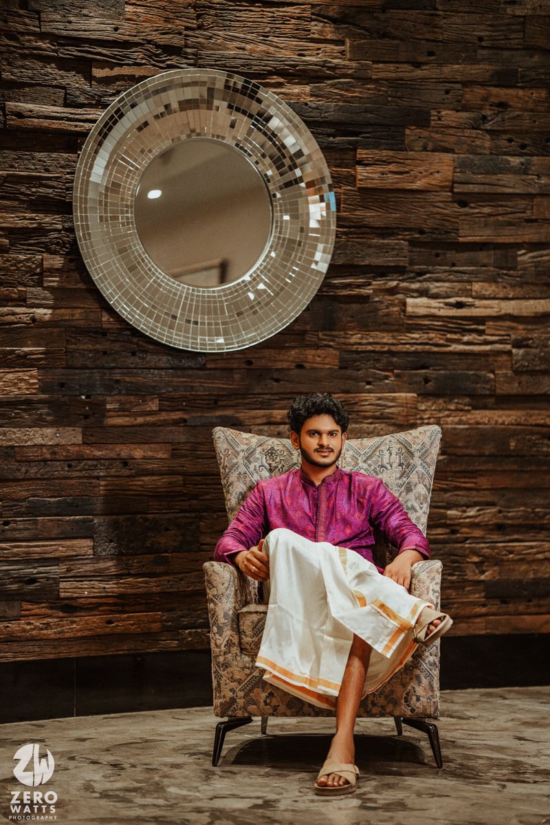 Young man in pink kurta and white dhoti seated in ornate chair against rustic wood wall with round mirror.
