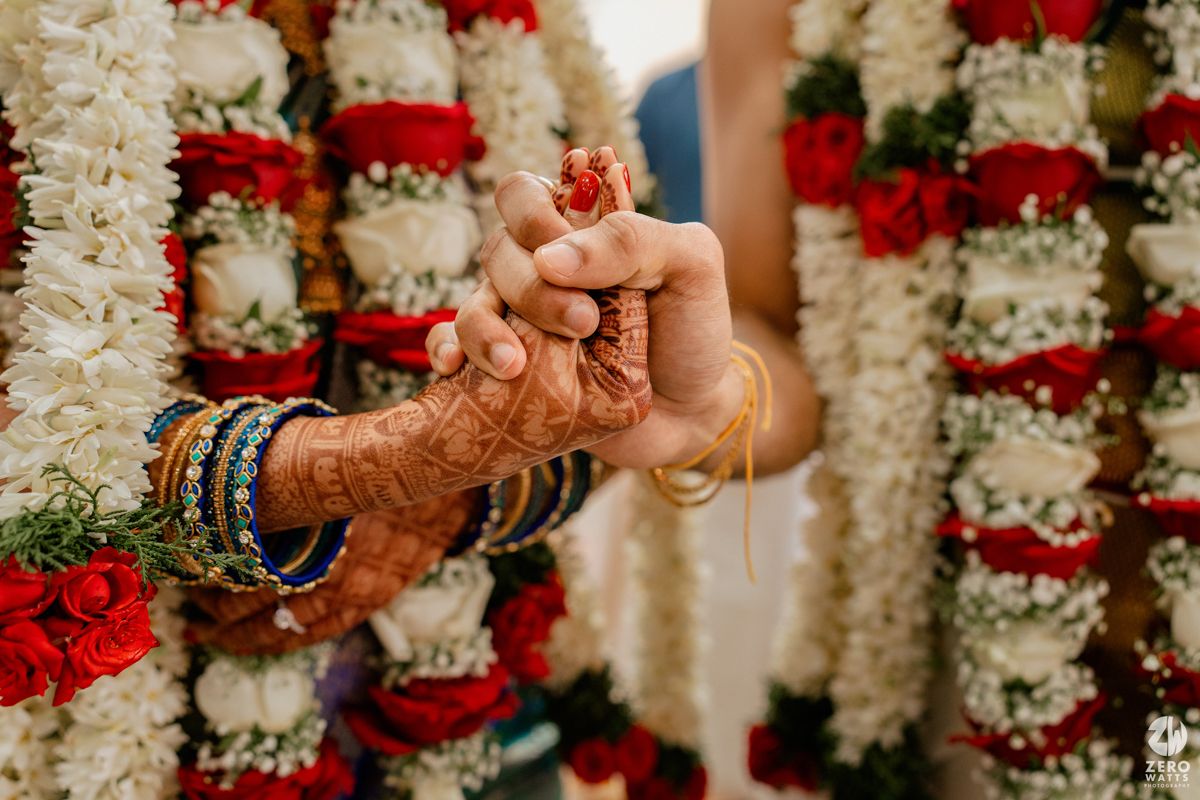 Close-up of bride and groom exchanging rings with intricate mehndi and bangles, surrounded by red and white floral garlands.