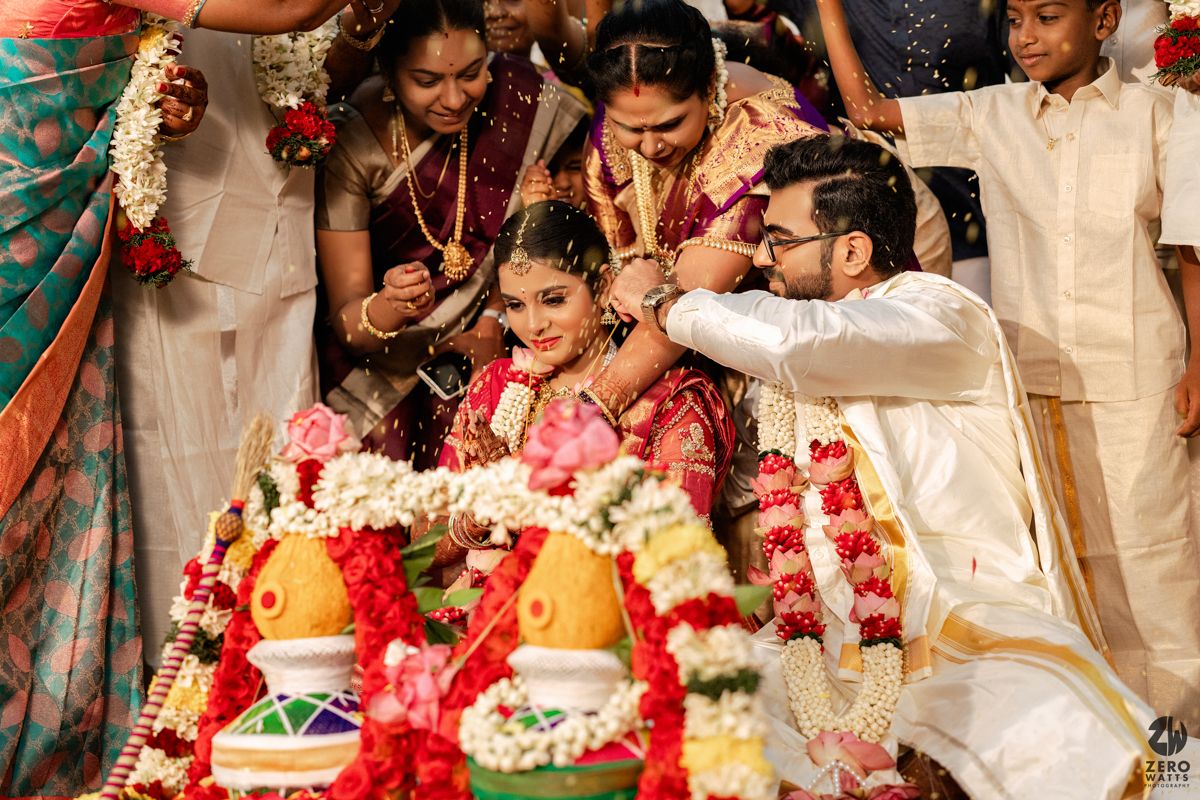 South Indian wedding ceremony with groom placing garland on bride surrounded by family and flowers.