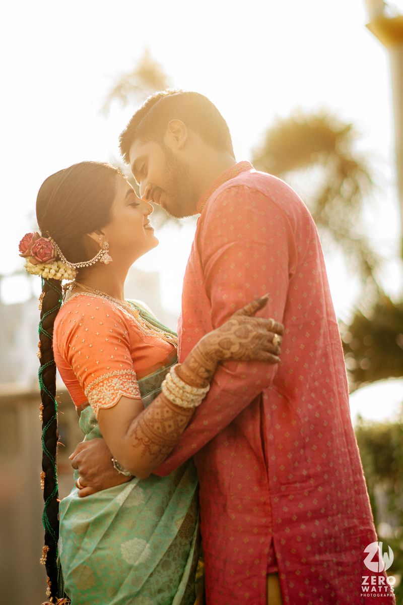 A couple in traditional Indian wedding attire share a romantic embrace at golden hour, bride in green saree and groom in pink kurta.