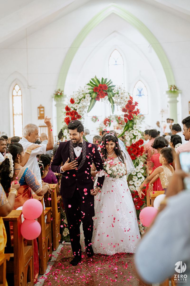 Bride and groom walk down church aisle showered with rose petals, guests cheering beside floral arch decoration.
