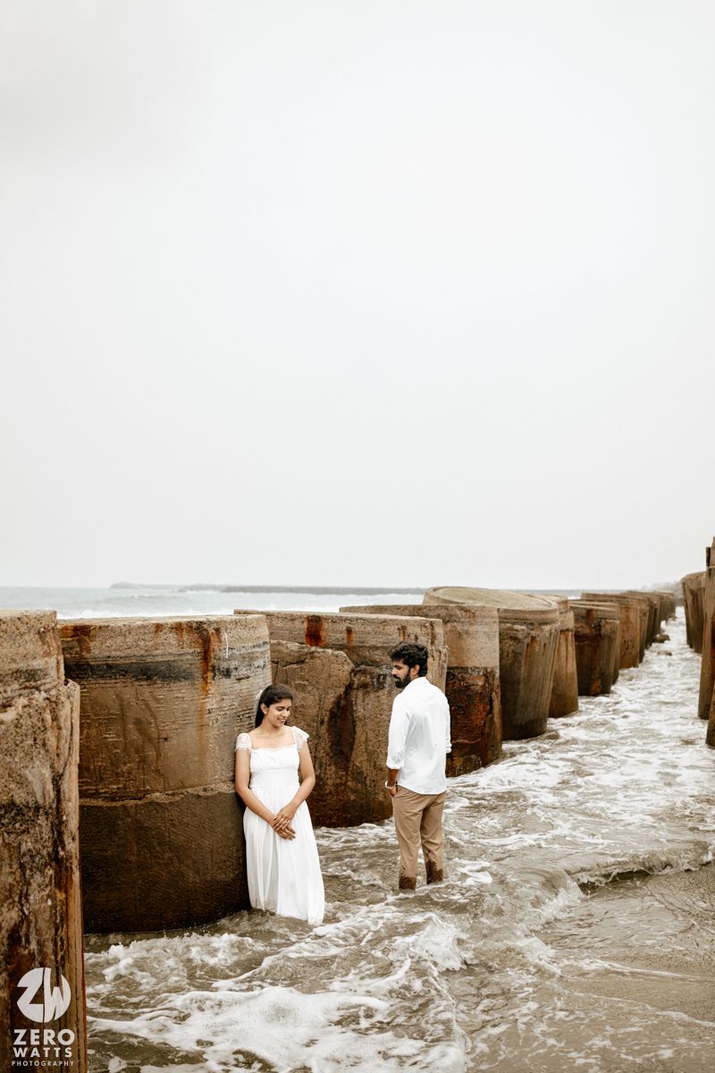 Couple in white outfits standing between rustic ocean pier pillars with waves washing around their feet.