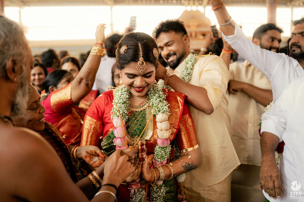 South Indian wedding ceremony with bride in red Kanjivaram saree receiving garland from groom amid joyful guests.