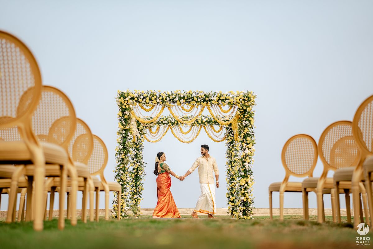 Indian bride and groom hold hands under a white and yellow floral mandap at an elegant outdoor wedding.