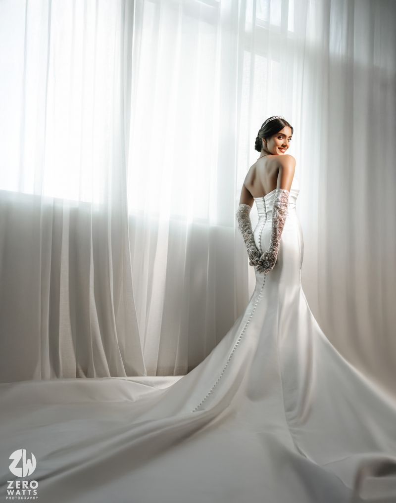 Bride in elegant white mermaid wedding gown with lace gloves and long train poses by sheer curtains.
