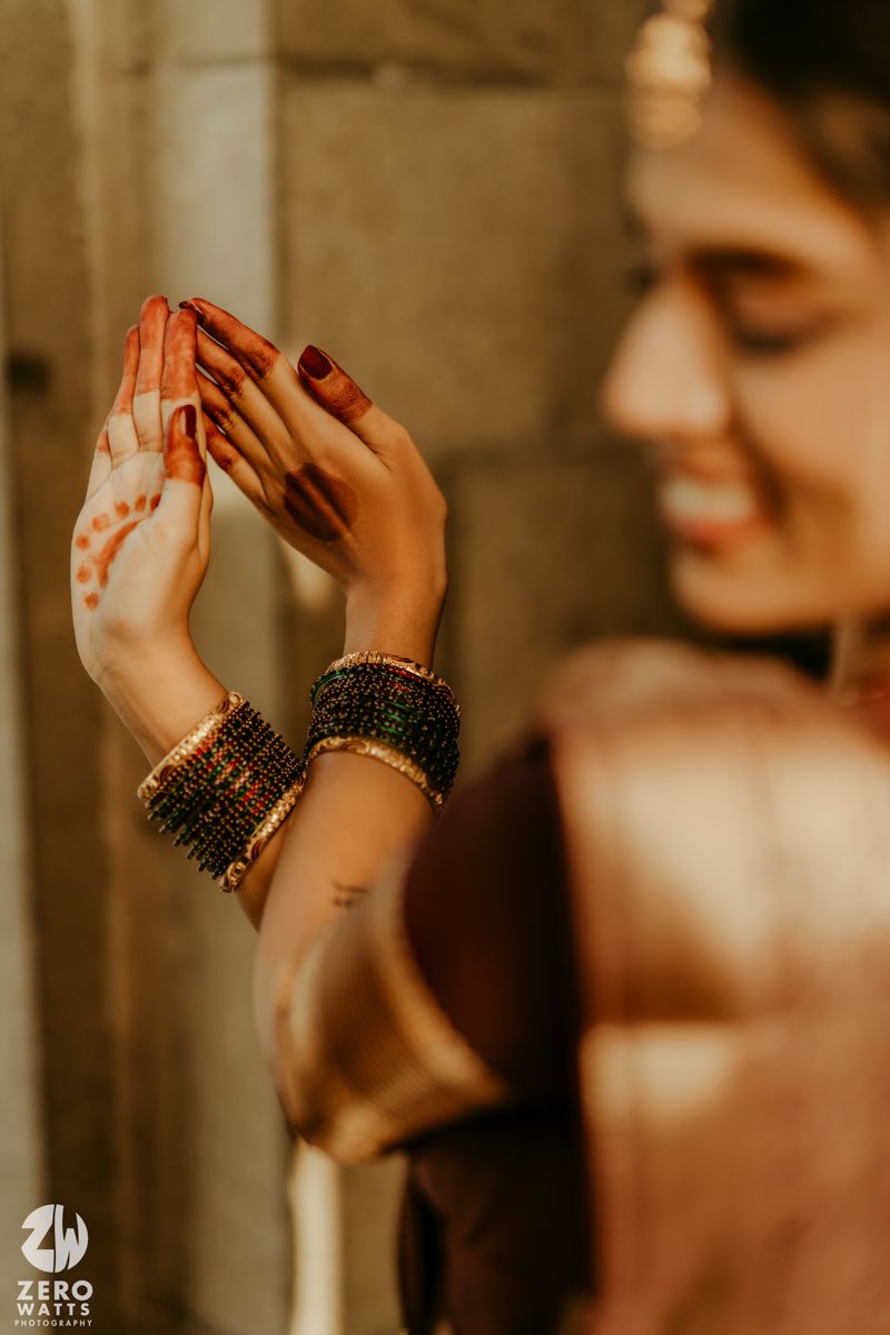 Bride showcasing intricate mehndi henna designs on hands with gold and dark bangles in a brown silk saree.