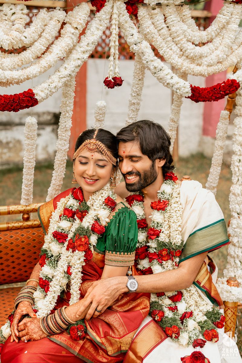 South Indian bride and groom in red silk saree and white dhoti adorned with floral garlands at wedding ceremony.