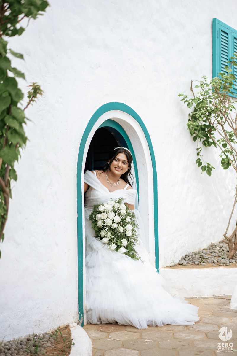 Smiling bride in white off-shoulder gown holds white bouquet standing in teal-trimmed arched doorway of white building.