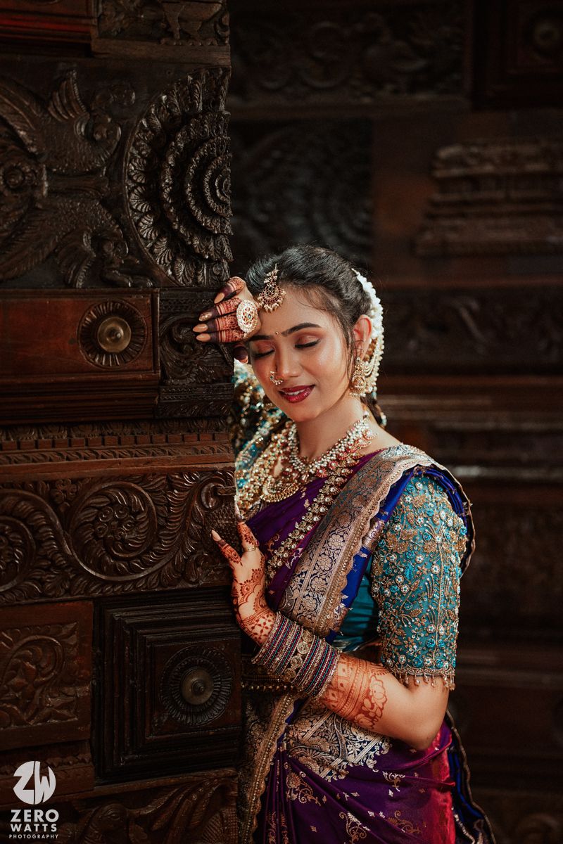 Indian bride in purple silk saree and gold jewelry poses gracefully by ornate carved wooden door.