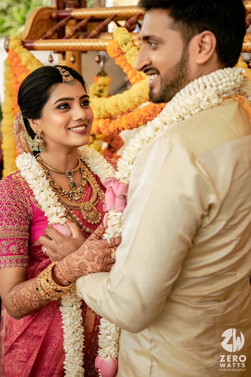 South Indian bride in pink saree with gold jewelry exchanges garlands with groom during traditional Hindu wedding ceremony.
