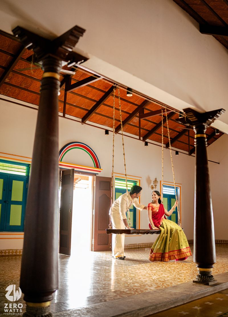 Bride in green and red saree sits on wooden swing as groom pushes her in a traditional South Indian home.
