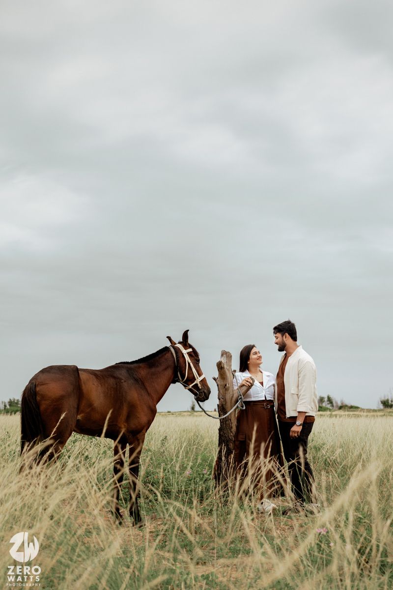 A couple stands together in a grassy meadow with several brown horses grazing around them.