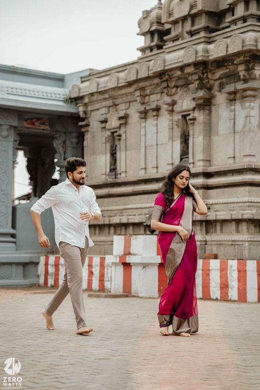 A couple in pink and white outfits dances playfully in front of an ornate temple structure.