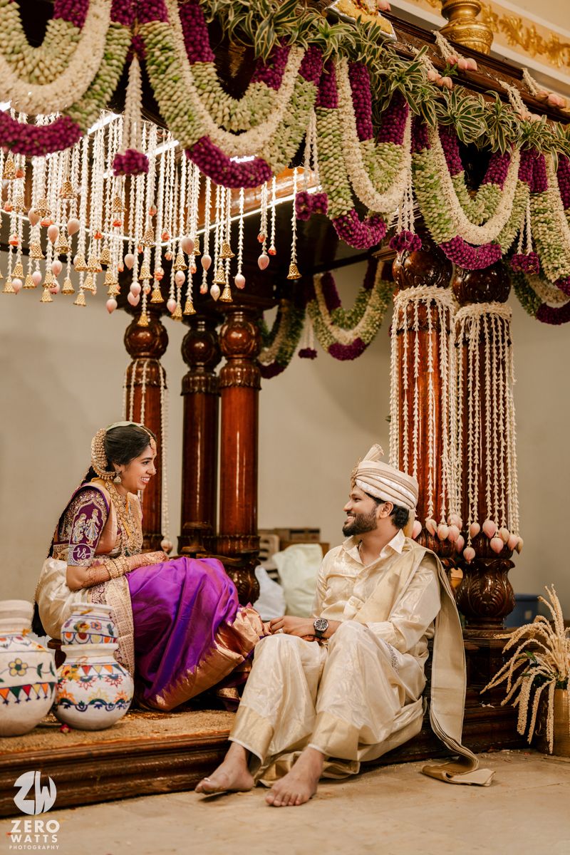 Couple in traditional Indian attire seated on floor during wedding ceremony with hanging floral decorations above.