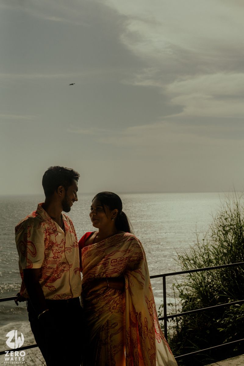 A couple in traditional Indian wedding attire stands against an oceanfront backdrop at dusk.