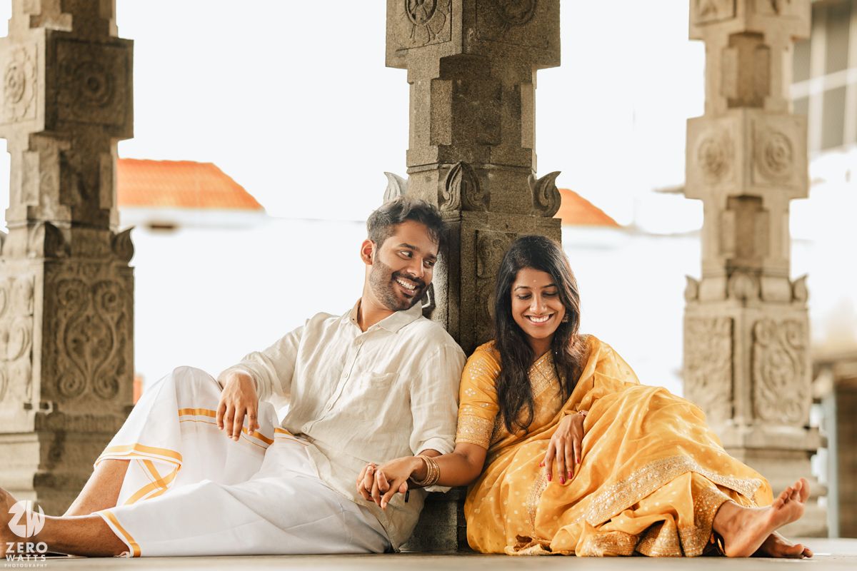 A couple in traditional Indian attire sits and laughs together among ornate stone pillars in an ancient temple.