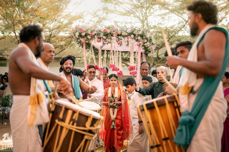Traditional Indian wedding procession with drummers in white dhotis accompanying a bride in red attire outdoors.