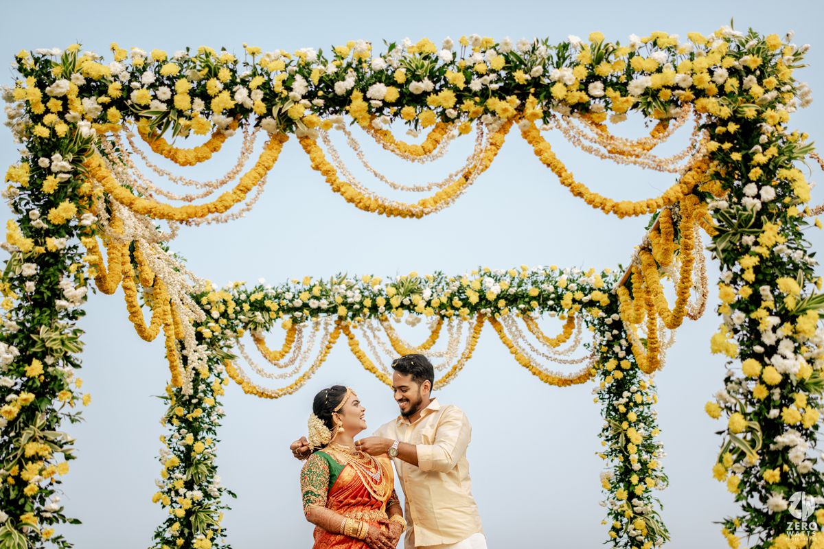 Indian couple poses joyfully under a floral mandap adorned with yellow roses and marigold garlands at their wedding.