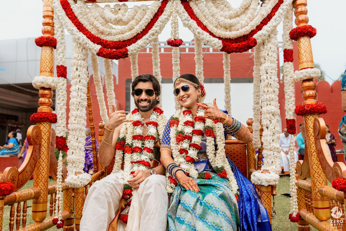 Indian couple in traditional wedding attire seated under floral mandap, smiling joyfully during vibrant Hindu wedding ceremony.