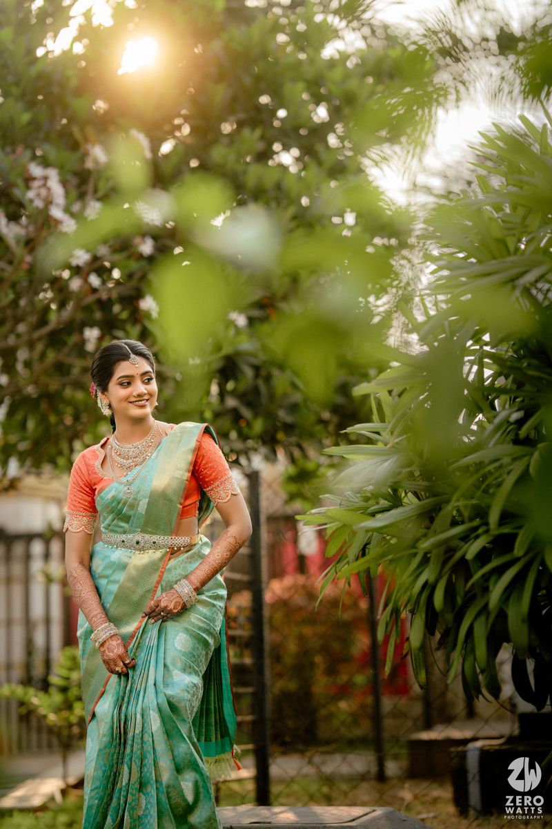 A bride in a teal silk saree with orange blouse and gold jewelry poses gracefully among lush green garden foliage.