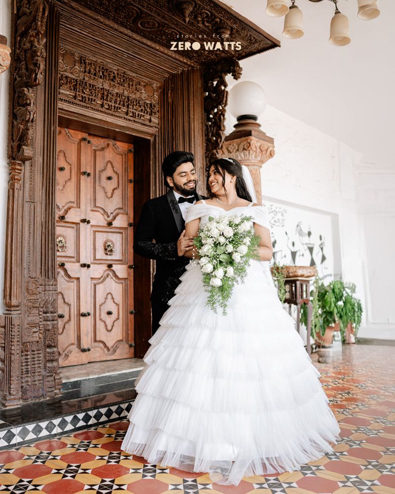 Bride in white ruffled gown holds bouquet with groom in black tuxedo before ornate wooden door on tiled floor.