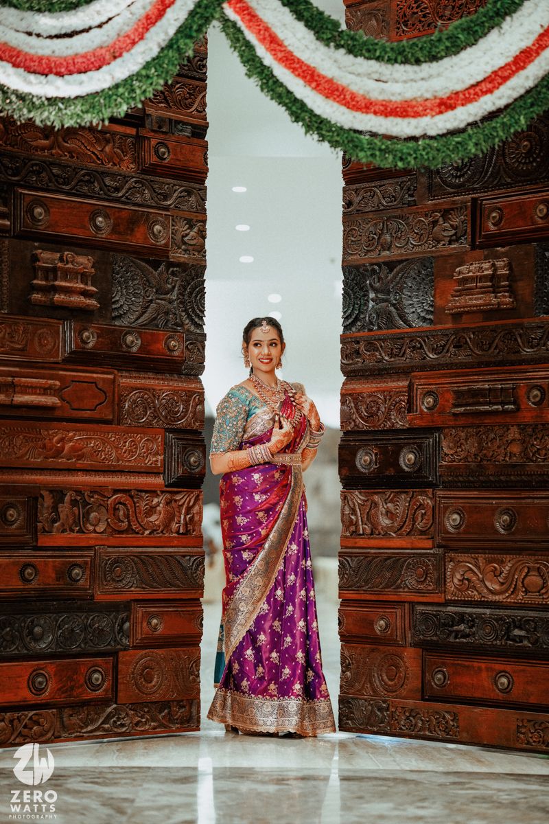 Indian bride in purple saree stands between ornate carved wooden doors adorned with floral garlands.
