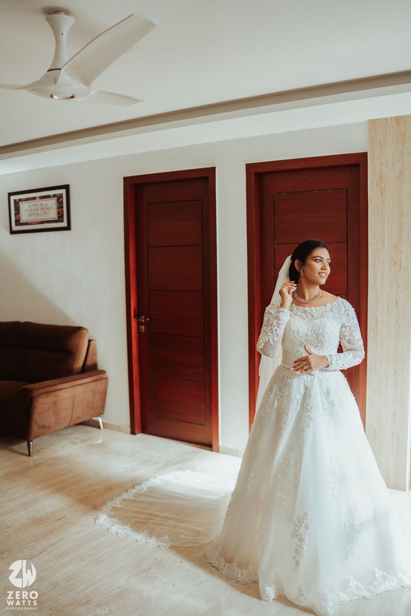 Elegant bride in long-sleeved lace wedding gown poses gracefully in front of wooden doors in a bright room.