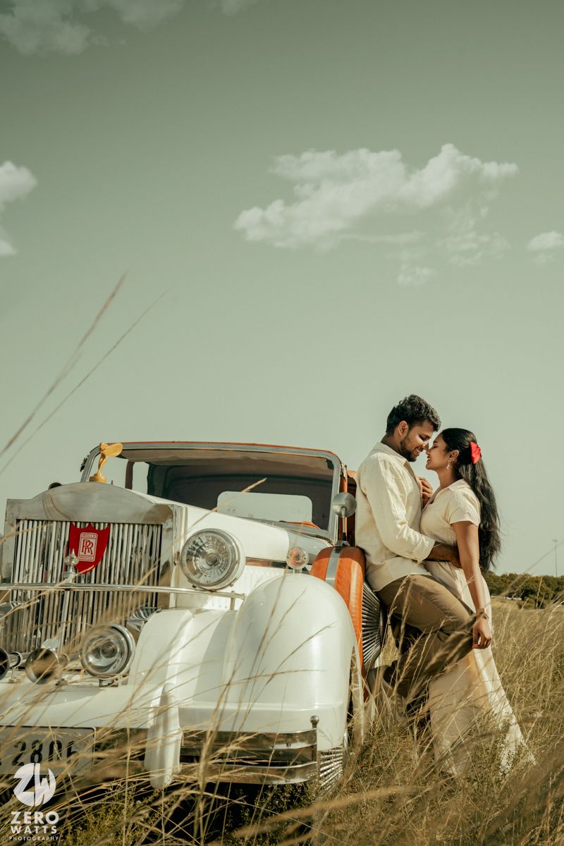 A romantic couple poses with a vintage white car in a rustic field setting with a dreamy green sky.