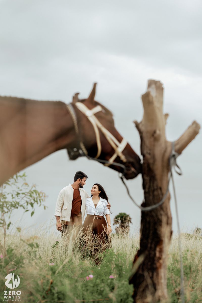 Romantic couple shares tender moments in a grassy field with horses on an overcast day.