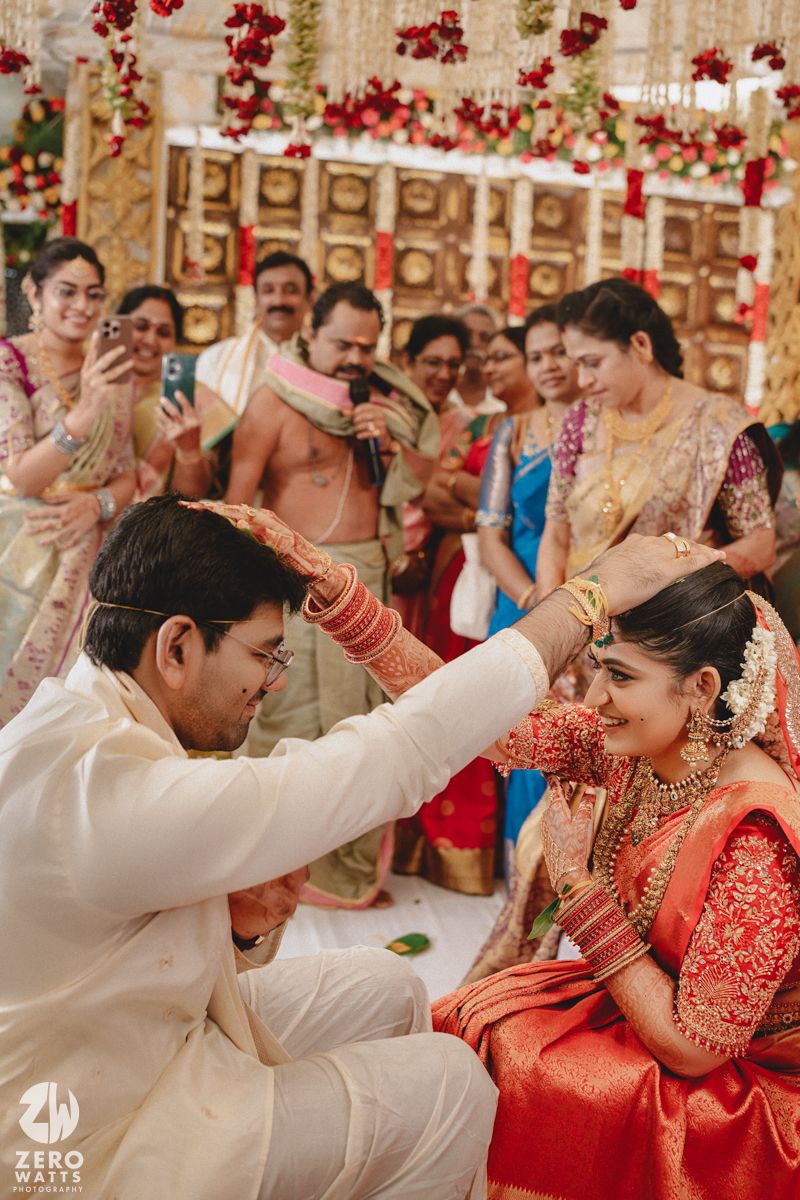 A traditional South Indian wedding ceremony with the couple performing rituals under decorative red floral arrangements.