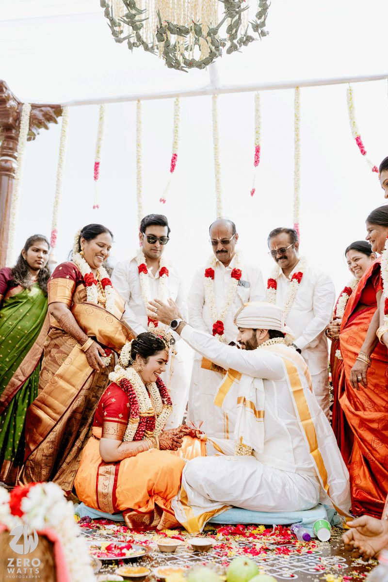 South Indian wedding ceremony with bride in red saree and groom in white performing traditional rituals under floral mandap.