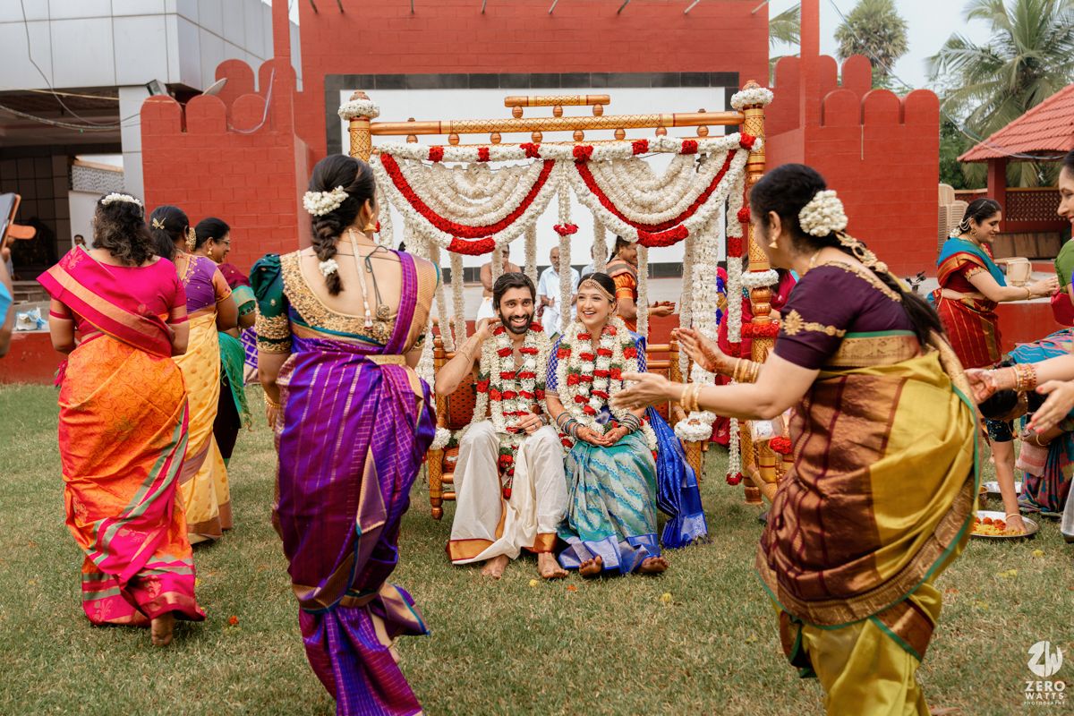 Colorful South Indian wedding ceremony outdoors with guests in vibrant sarees celebrating the bride and groom under a floral mandap.