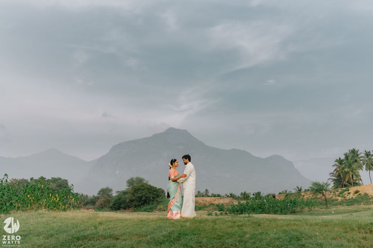 South Indian wedding couple embracing in a scenic green landscape with dramatic mountain backdrop and cloudy sky.