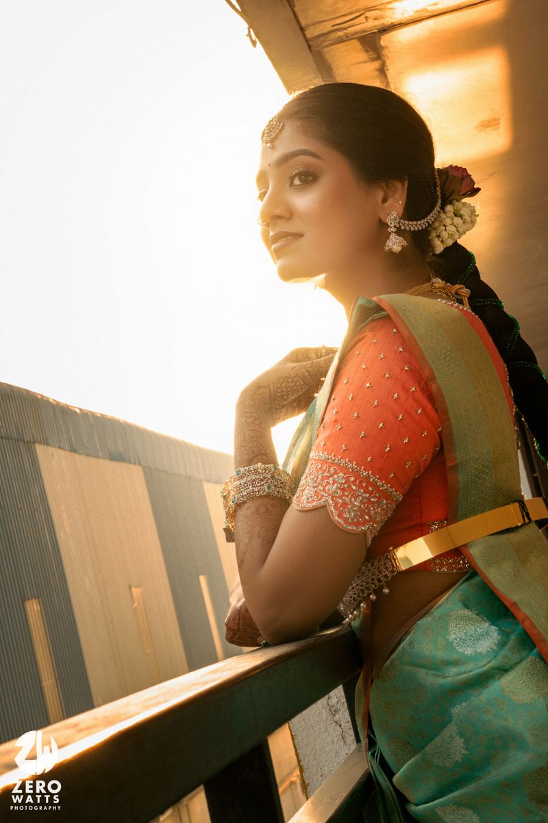 A bride in a teal and orange saree with gold jewelry poses on a balcony in warm sunlight.